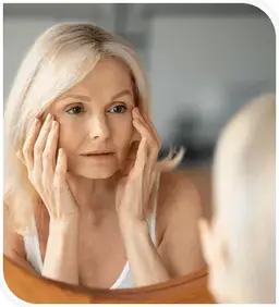 A woman in her fifties studying her reflection in a mirror, hands on her cheeks